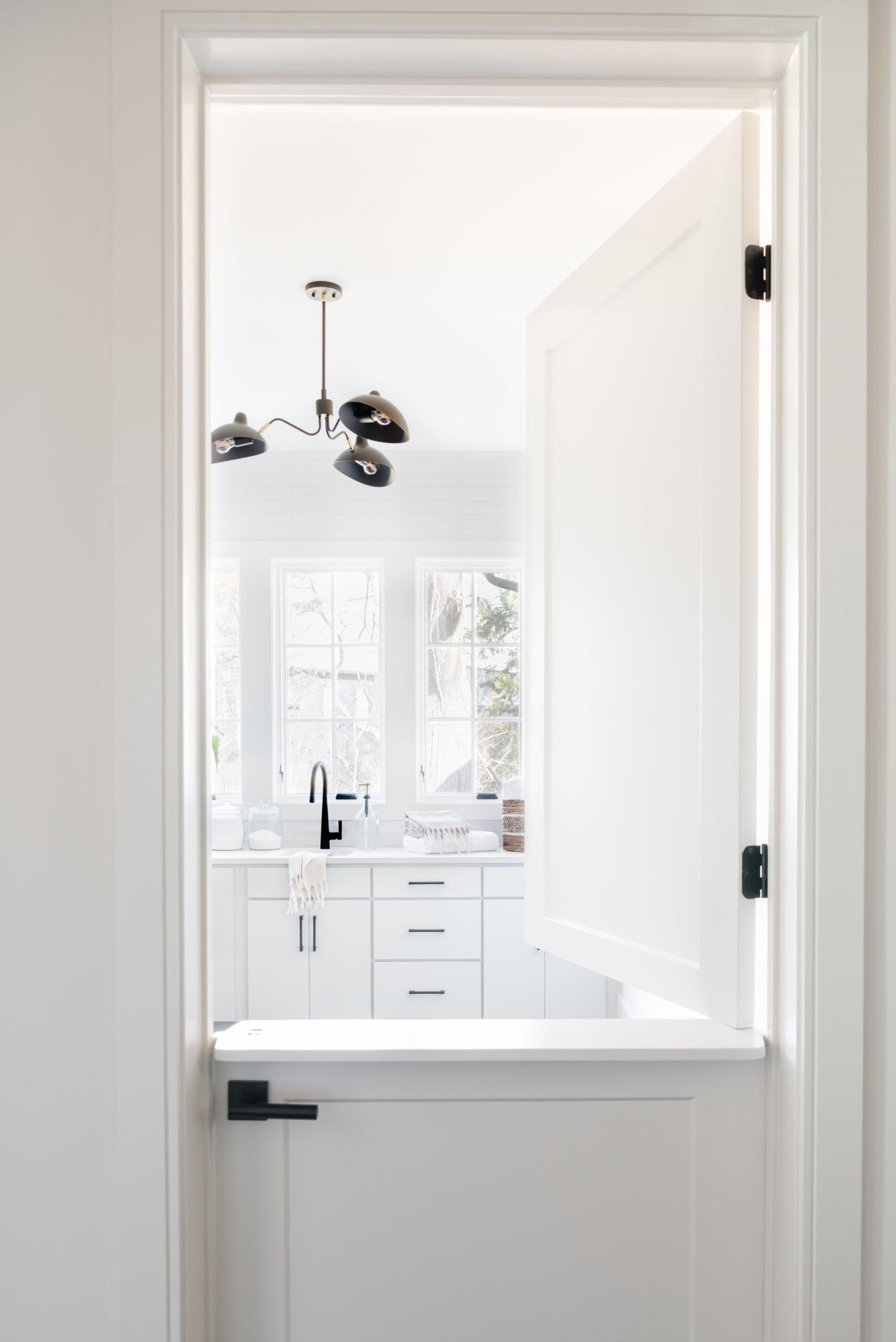 White dutchdoor leading into a white laundry room with window wall and black modern chandlier