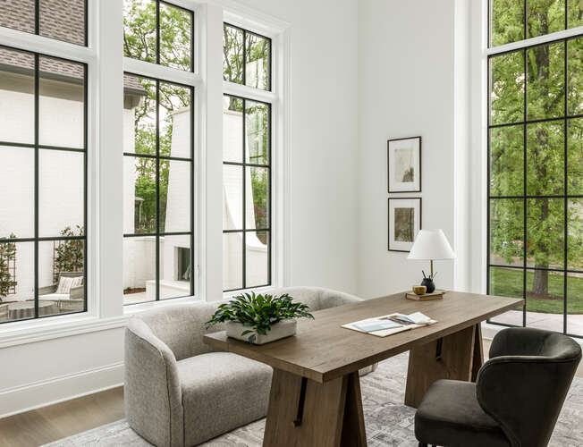 Modern office with three black grid tall windows, white walls, and a wood desk with modern upholstered chairs