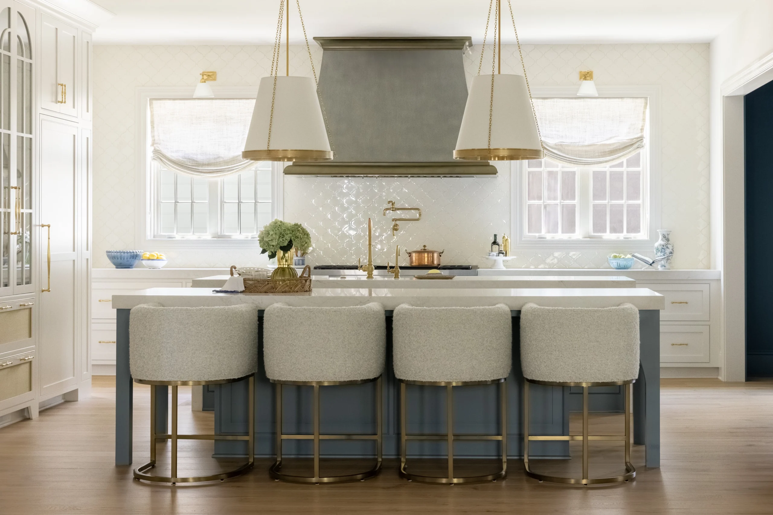 White kitchen with large island, gold pendants, upholstered stools, and two windows framing the range