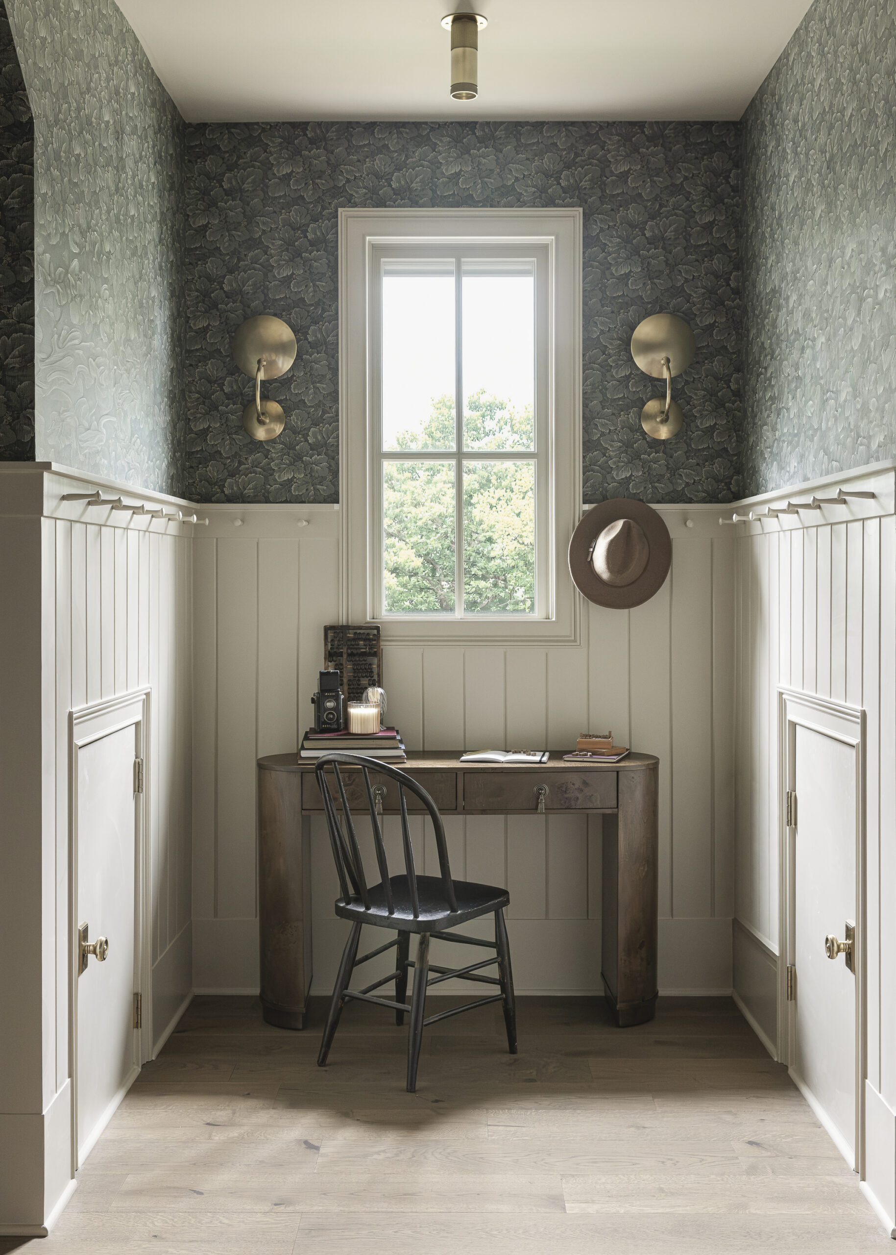 Small room with green wallpaper and creamy paneling complete with pegs for added hanging storage with two gold sconces for soft light framing a window