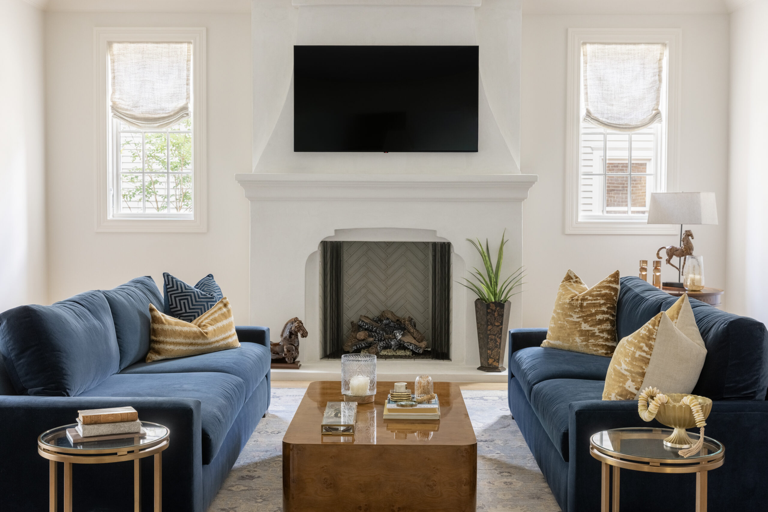 Family room with centered fireplace with white mantle and windows on each side and navy couches and modern wood coffee table