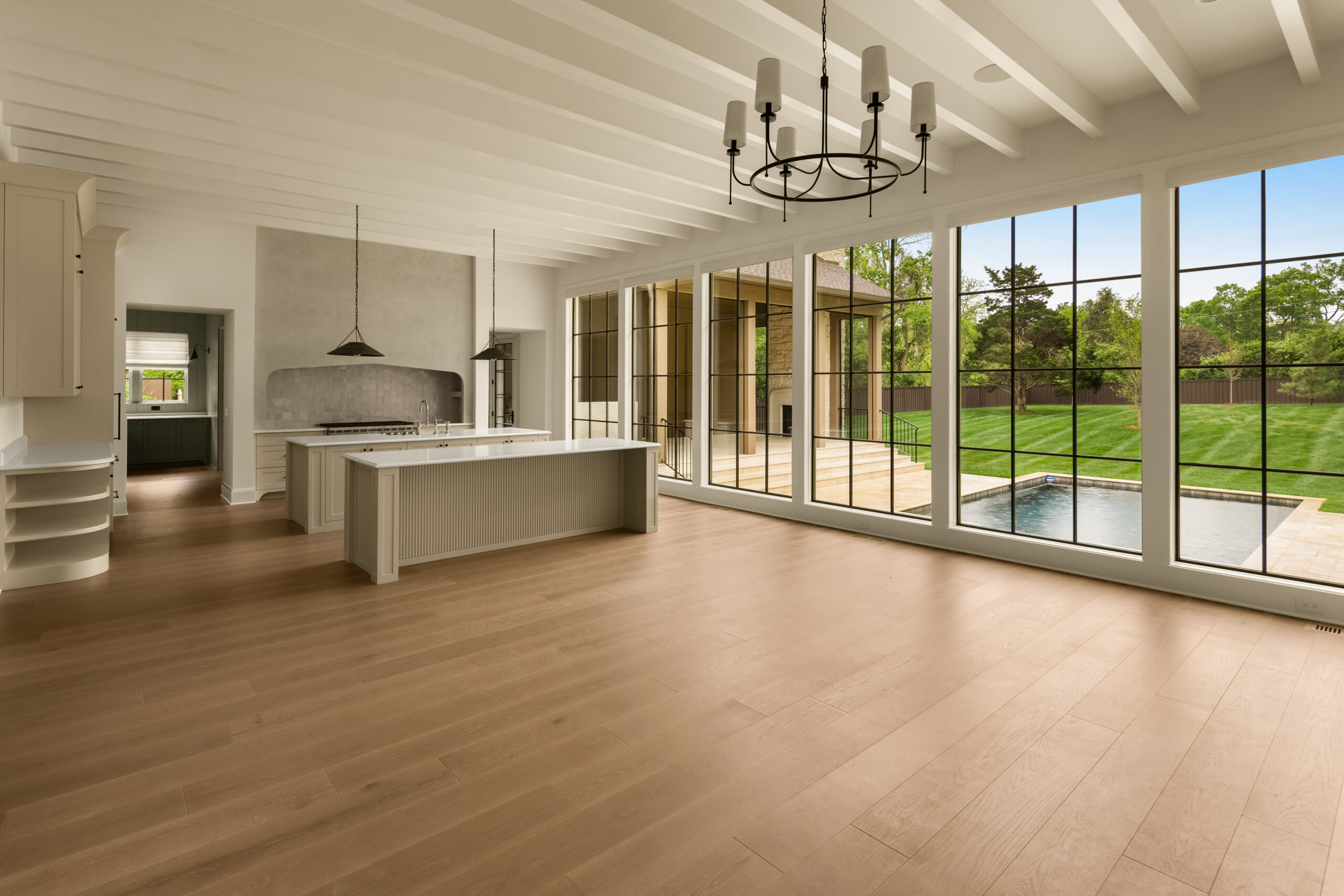 Creamy white walls with floor to ceiling large windows with black grids with a view of the fluted kitchen island and arched range hood, painted ceiling beams, and a circular black iron chandelier