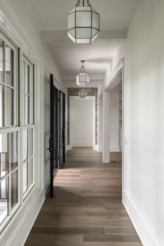 Hallway lined with windows to the left and white walls to the right with chandeliers spaced along the ceiling and brick walls framing the door at end of hall opening