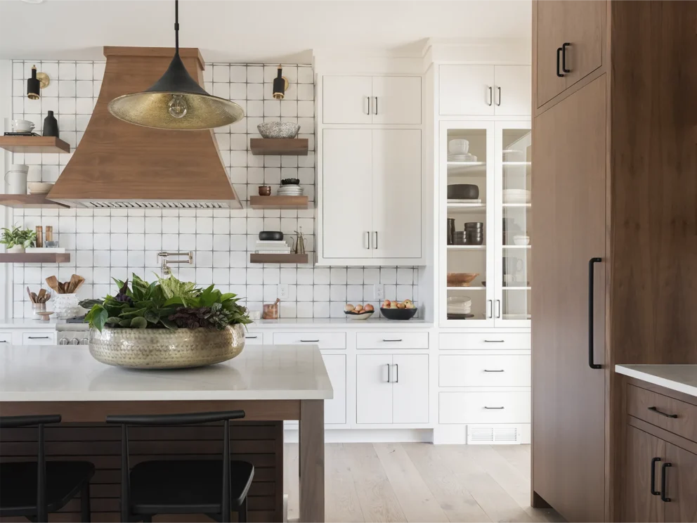 Bright modern kitchen with warm wood cabinetry, white cabinets, brass pendant, copper range hood, and a lush island plant creating a welcoming space
