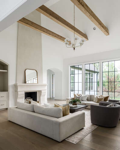 High ceiling family room with wood beams, white walls, taupe stucco fireplace and a wall of black grid windows