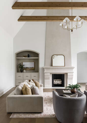 High ceiling family room with wood beams, white walls, taupe stucco fireplace, and arched open shelved built-ins